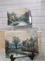 Two of the rectangular Thomas Kinkade collector plates with village brook and cottage scenes, shown on clear stands against a wood-panel background.