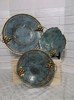 Photo showing three decorative plates with patina finish: two round plates with decorative brass grape clusters and one leaf-shaped plate also with brass grape cluster accents.