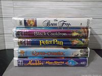 Front view of all five sealed Disney VHS tapes stacked horizontally showing titles on the spines: The Brave Frog, Black Cauldron, Peter Pan, Quest Camelot, Aladdin and the King of Thieves.