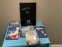 Photo showing five children's DVDs on a blue cloth, with a black folder in background marked 'Lot #31'. Titles visible include Finding Nemo, Maleficent, Ghostbusters 35th Anniversary, Monsters Inc., and Quest for Camelot.