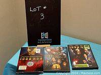 Three DVD cases placed upright on a blue surface with a black sign behind them. Leftmost DVD is Iron Man 1, center is Iron Man 2, rightmost is also Iron Man. Condition varies: one unopened disc edition, two opened cases.