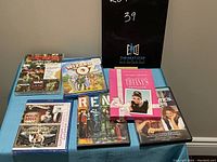 Full view of seven musical DVDs on a blue cloth surface with black sign in background showing lot number