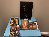 Four DVD and Blu-ray movie cases placed on a blue surface with a black folder behind them. Titles include National Treasure, The Da Vinci Code Trilogy, The Musketeer, and The Order.