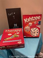 Two boxed games Therapy and Yahtzee on blue cloth surface near a black backdrop with lot number written.