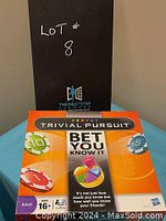 Photo of the closed front of the Trivial Pursuit Bet You Know It game box sitting on a table with a sign behind it indicating lot number 8.