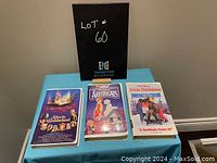 Front view of three VHS tapes with titles: Alice in Wonderland, The Aristocats, Cool Runnings, displayed on a blue cloth with a black auction sign behind them.