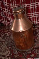 Copper milk jug standing upright on a patterned fabric surface with a red and white checked background. The jug has a lid with a chain, handle, and stamped letters on its surface.