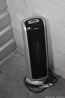 Single photo showing the Lasko oscillating tower heater placed against a concrete block wall. Heater is silver and black with a front mesh grill and control panel on top.