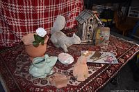 Various pottery pots, planters including a frog planter, a wooden birdhouse, squirrel figurine, decorative tiles and other outdoor decor on a patterned rug and red plaid backdrop.