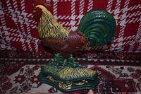 Photo shows the painted cast iron rooster figurine on a patterned carpet with a red plaid or checkered background. The rooster is painted in multiple colors and mounted on a green base with gold trim.