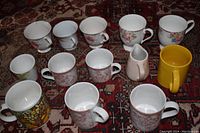 Twelve porcelain coffee cups with various floral and colorful designs arranged on carpet, one yellow cup and one pink creamer pitcher.