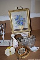 Photo of a vintage small easel holding a gold-framed floral print, surrounded by various small decorative objects including glass cups, a vase, two small framed images, a porcelain creamer, a decorative apple, and a small plate.