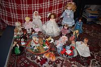 Full collection of 23 vintage dolls of varying sizes and styles photographed together on a patterned rug in front of a red and white plaid background.