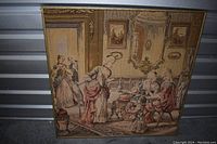 Photo of the vintage framed tapestry with a gold gilded frame showing an 18th-century style social scene of ladies sipping wine, with some visible minor damage to the frame.