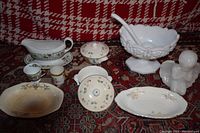 Lot of 10 vintage serving pieces on red and white patterned blanket, includes white milk glass punch bowl, porcelain gravy boat, floral serving bowls, cups, lidded bowl, and angel figurine