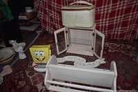 Photo showing white wooden storage box with open glass doors and glass shelf inside, cream vintage-style carry-on bag on top, white wooden wall shelf in front, yellow SpongeBob container, ceramic dish, small angel figurine, curved wooden signs