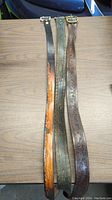 Three leather belts laid flat on a wood-grain surface, showing full length and metal buckles