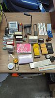 Overview of desk items including calculators, stamps, staplers, and other office supplies arranged on a wood grain desk surface.