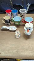 Overview photo showing assortment of Disney-themed mugs, plastic cups, ceramic spoon, small figurine, and Olaf cup on a wooden surface.