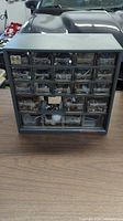Front view of black plastic storage cabinet with multiple clear drawers containing assorted metal hardware pieces, placed on a wood surface.