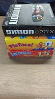 Stacked box set showing three games: Simon Optix on top, Pie Face middle, Hedbanz bottom on a wood surface