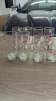 Set of 8 clear Coca-Cola glasses with various Christmas-themed decorations including Santa in red and white suit and colorful holiday houses on a wooden table.
