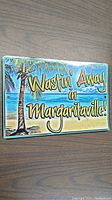 Metal rectangular sign lying flat on a wood surface showing a colorful tropical beach scene with a palm tree and the text 'Wastin' Away in Margaritaville!'