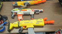Six Nerf blasters displayed on a table in two rows. Top row: yellow/gray, white/orange, yellow 'FORTNITE'. Bottom row: orange/gray, blue, gray/blue.