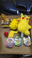 Lot items arranged on table showing Pikachu plush toys, Pokémon Digivices in collectible tins, golden figure, and Pokéball.