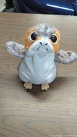 Photo shows a Star Wars Porg plush toy on a table, featuring its orange, gray, and white coloring and large eyes.