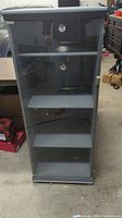 Full front view of gray stereo component cabinet with glass door closed, showing three shelves and slats for ventilation on the back panel.