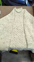 Full view of cream-colored cable knit sweater laid flat on table, showing full front and sleeves with textured knitting and speckled yarn.