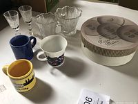 Shot showing 3 ceramic mugs, 2 sherry glasses, 2 cut glass dishes, and the round petit four plate box on a white surface