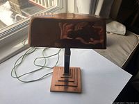 Front and side view of vintage copper bankers lamp on white surface near window showing shade and stepped base details.