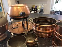 Overview of the lot showing copper fondue pot with stand and burner, a ribbed copper bucket, two small copper cups, and smaller pot with lid and spout on a countertop.