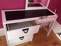 White wooden desk with a black surface on top, two drawers with black handles on bottom drawer and missing on top. Mirror frame attached at desk back.