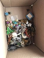 Photo of a cardboard box filled with various vintage costume brooches and clip-on earrings, showing a mix of colorful and metal pieces with rhinestones and natural stone accents.