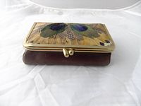Closed small brown leather case with brass frame and peacock feather lid decoration, sitting on white background.