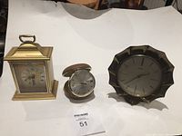 Three vintage clocks displayed on a white surface: brass petal-shaped clock, Tempus Fugit mantel clock, small silver-tone travel alarm clock.