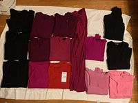 Top-down view of 15 folded women's cashmere sweaters in various colors including black, burgundy, maroon, fuchsia, pink, and orange laid on a white sheet on wood floor.