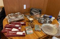 Overall view of lot on wooden table including 12-section serving tray, napkin rings in boxes and loose, metal baskets, silver cleaning supplies, and serving utensils.