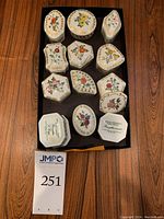 Photo shows all 12 porcelain pill boxes with varied floral designs and their shapes laid out on dark tray.