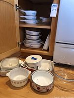 Photo showing Pyrex Cornflower bakeware and casserole dishes stacked inside wooden cabinet, plus an orange Cerutil stoneware pan and small Corning Ware pots and pans with lids on counter.