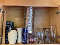 Photo shows nine various vases in a wooden cabinet including porcelain, cut glass, and crystal varieties in different colors and heights.