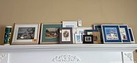 Wide angle photo showing the collection of framed and unframed wall art arranged on a mantel shelf, including a tall cylindrical container on the left.
