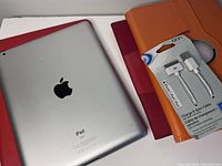 Apple iPad backside showing in silver with Apple logo and part of three folio cases and packaged charging cable.
