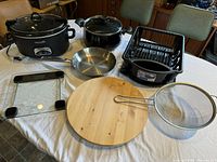 Full lot view on table showing all items: Crock Pot, frying pan, dish rack, digital scale, wooden lazy Susan and strainer.
