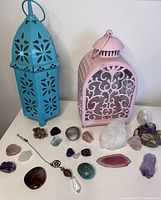 Photo showing two decorative lanterns (one turquoise hexagonal and one pink with scrollwork) alongside an assortment of various crystals, stones, and a pendant necklace arranged on a white surface.
