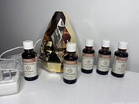 Full view of faceted gold Saje Aromagem diffuser with five Aveda essential oil bottles arranged around it, showing white caps and labels.