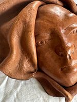 Close-up photo of the brown leather mask showing the molded face with detailed eyes, nose, lips, and ruffled leather edges.